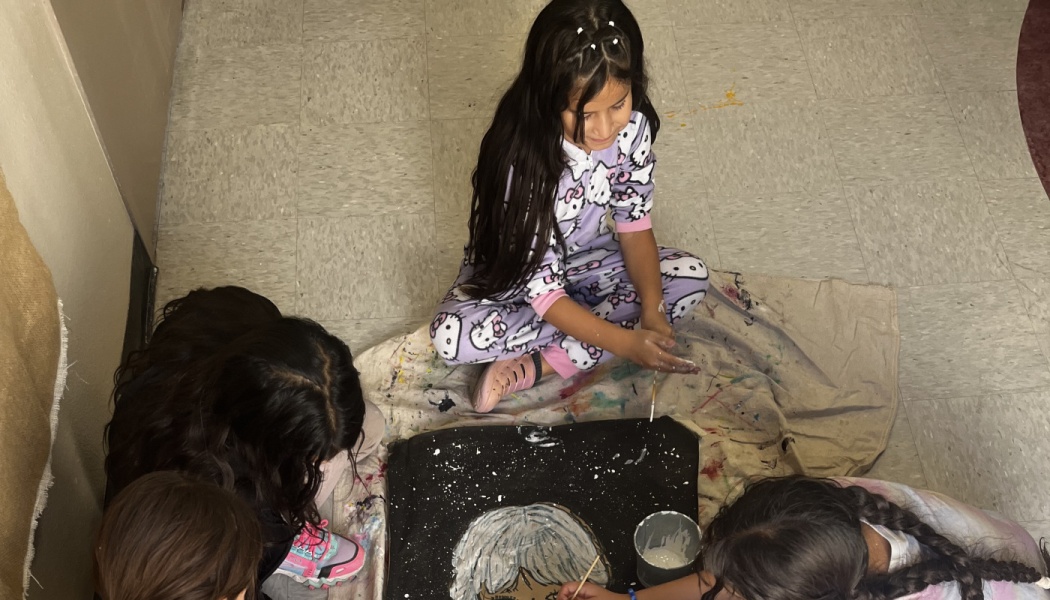 Young girls sitting on the floor together, collaborating on a canvas painting project.