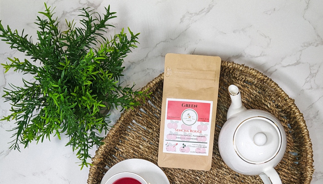 A pouch of Sencha Rouge tea, a white ceramic teapot, and teacup arranged on a woven tray next to a green plant.