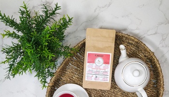 A pouch of Sencha Rouge tea, a white ceramic teapot, and teacup arranged on a woven tray next to a green plant.