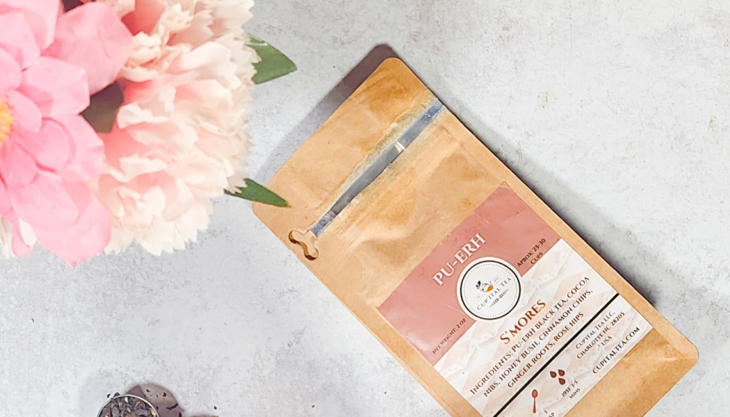 Kraft paper bag of Cupital Tea S'Mores Pu-Erh tea alongside soft pink flowers on a light gray surface.