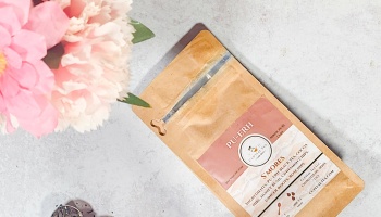 Kraft paper bag of Cupital Tea S'Mores Pu-Erh tea alongside soft pink flowers on a light gray surface.