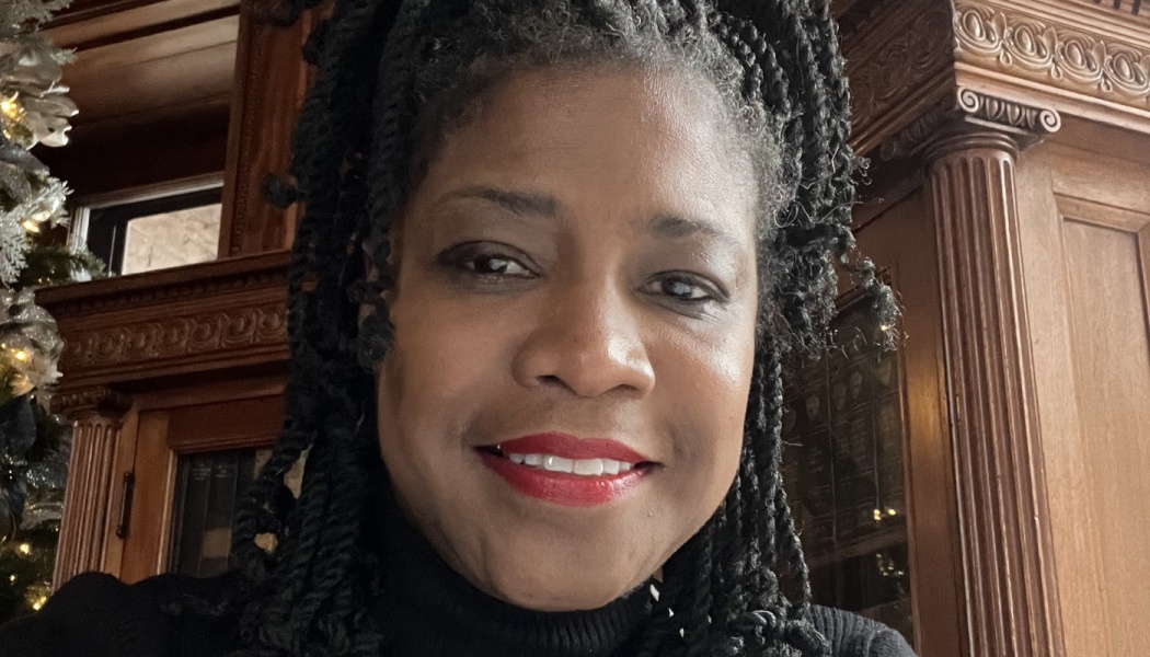 A smiling Black woman with dark twisted braids and red lipstick in front of ornate wooden paneling.