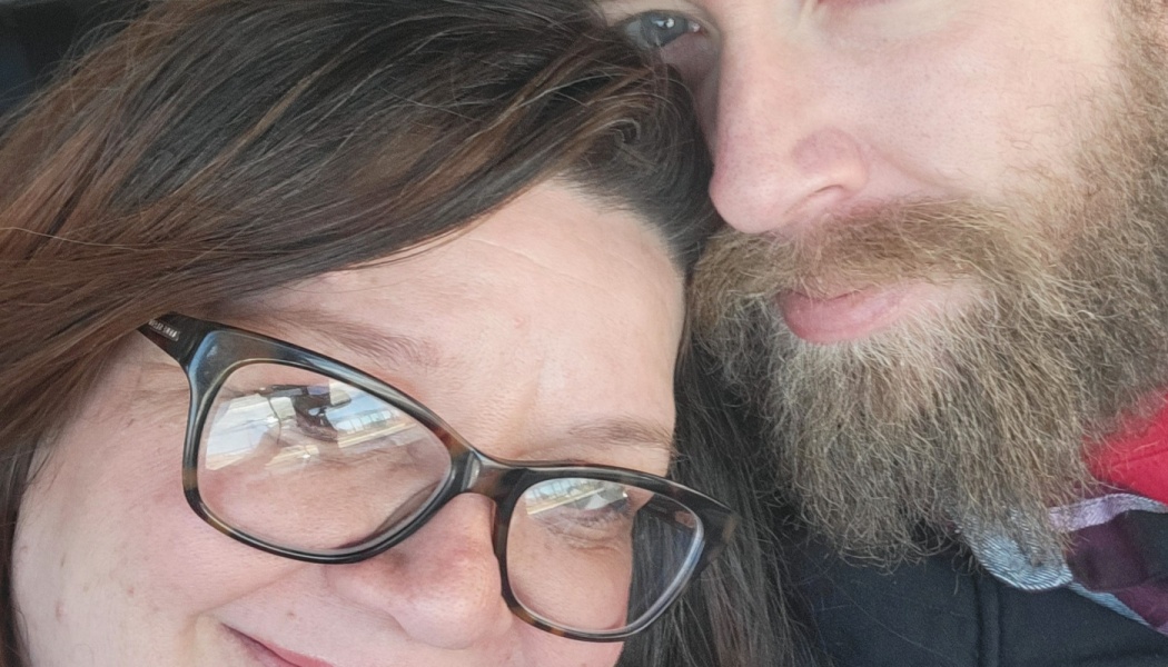 Close-up selfie of a smiling woman with eyeglasses leaning her head against a bearded man.