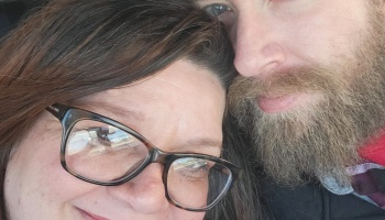 Close-up selfie of a smiling woman with eyeglasses leaning her head against a bearded man.