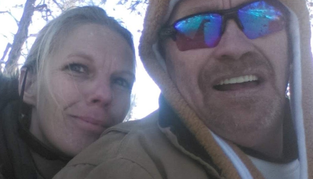 Close-up selfie of a smiling couple outdoors in hooded jackets, the man wearing reflective sunglasses.