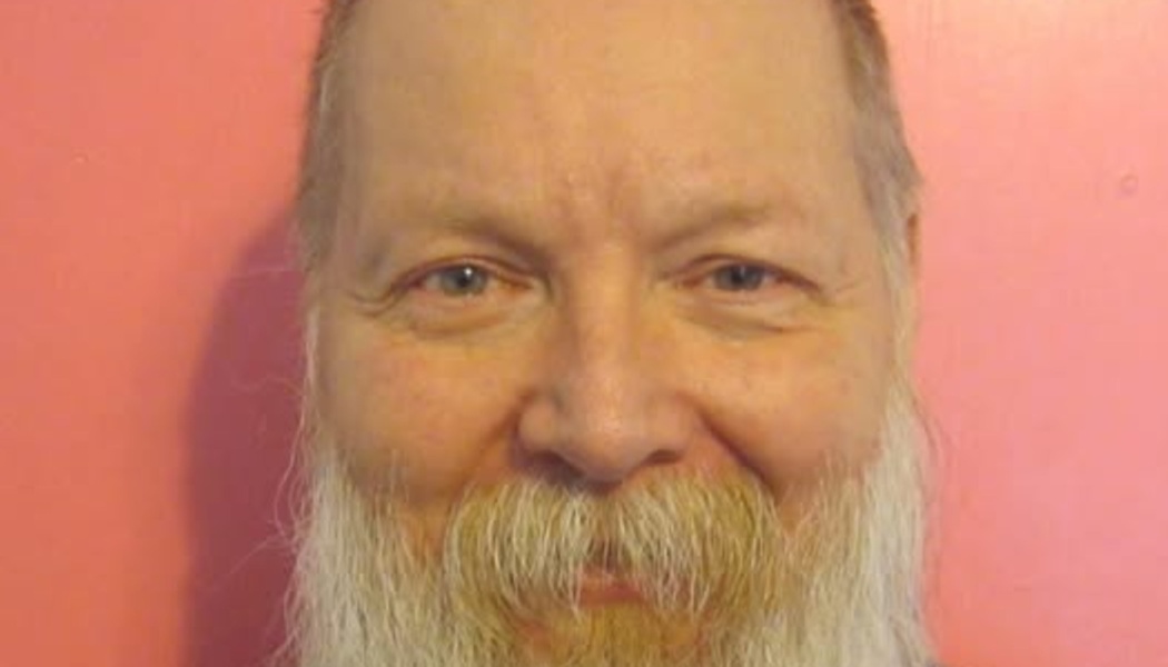 Close-up portrait of a man with a full white beard and blue eyes smiling against a pink background.