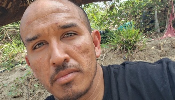 Close-up selfie of a man with a shaved head and goatee wearing a black shirt outdoors near greenery.