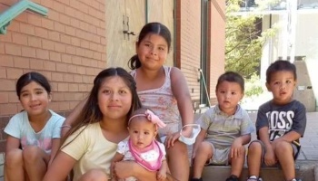 Six young children, including a baby, sitting together outdoors on steps near a brick building.