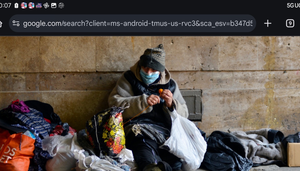A person wearing a face mask and beanie sits against a stone wall surrounded by bags and belongings, holding yarn.