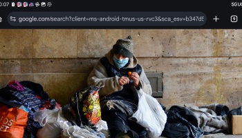 A person wearing a face mask and beanie sits against a stone wall surrounded by bags and belongings, holding yarn.