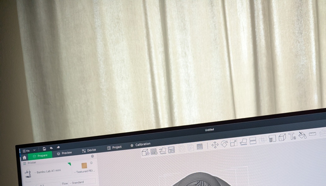 A computer screen displaying 3D printing software with a 3D model, set in front of cream curtains.