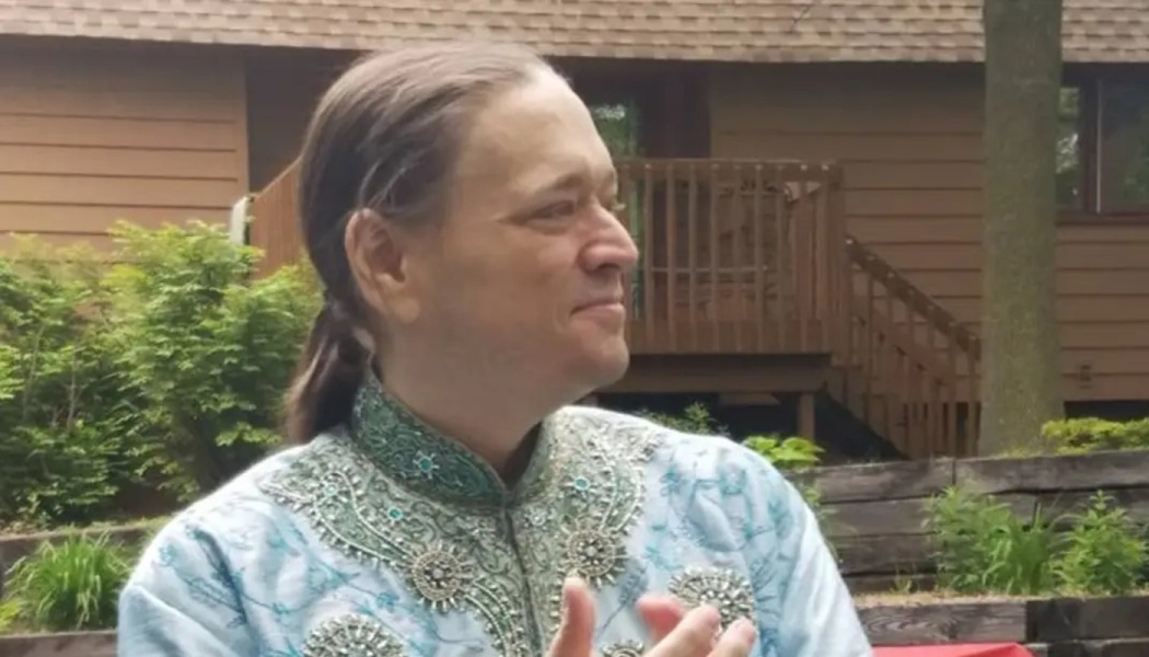 A smiling man with a ponytail wearing an ornate light blue embroidered tunic outdoors.