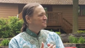 A smiling man with a ponytail wearing an ornate light blue embroidered tunic outdoors.