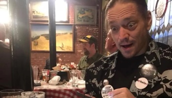 Wide-eyed man with a shocked, excited expression wearing a patterned jacket with pin buttons in a restaurant.