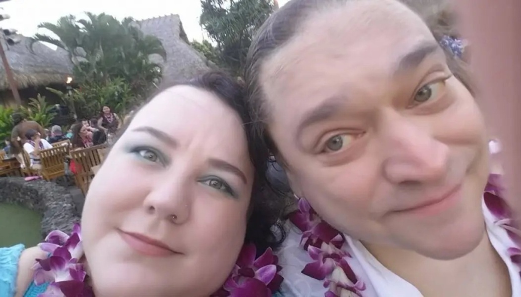 A smiling couple wearing purple orchid leis poses for a close-up selfie at a tropical resort.