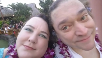 A smiling couple wearing purple orchid leis poses for a close-up selfie at a tropical resort.