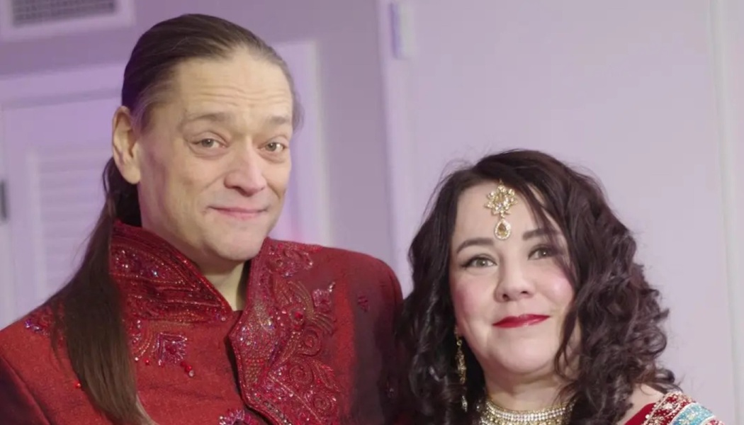 A smiling couple dressed in ornate, embroidered red traditional Indian attire and gold jewelry.