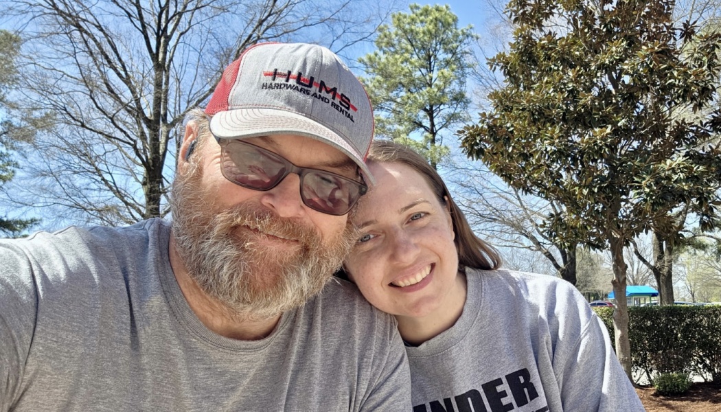 A smiling bearded man in a cap and glasses with a woman leaning her head on his shoulder outdoors.