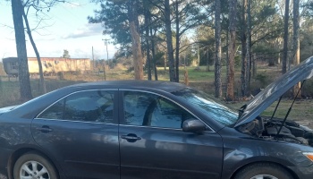 Dark gray sedan with its hood popped open, parked in a wooded rural area.