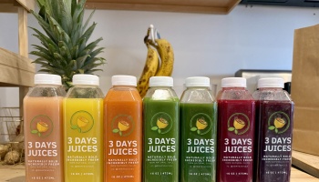 Seven colorful bottles of 3 Days Juices lined up with fresh pineapple and bananas in the background.