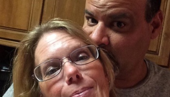 Close-up selfie of a smiling woman with glasses and a man leaning in close behind her.