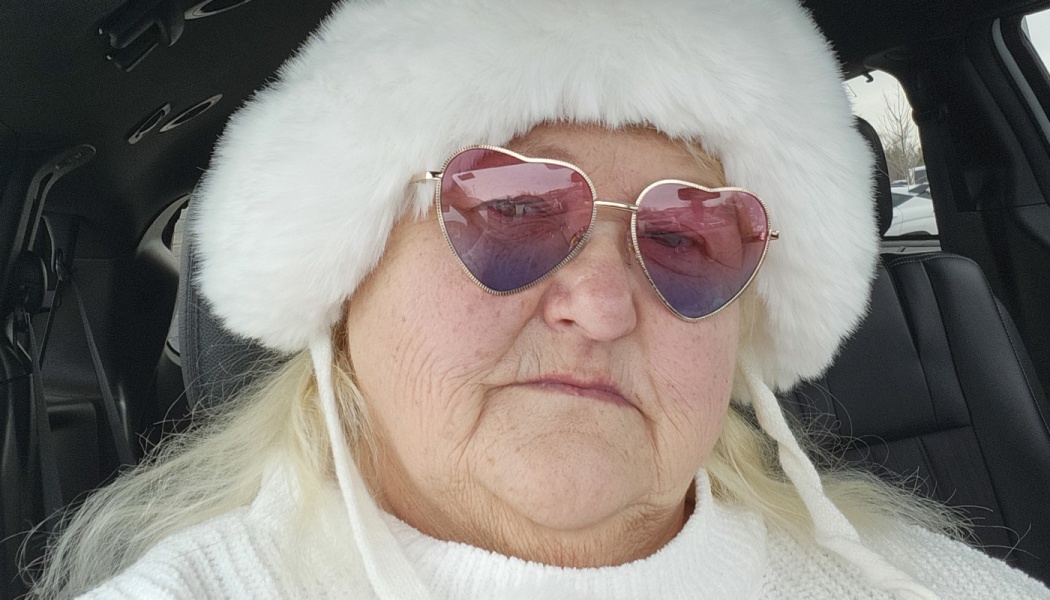 An older woman wearing a plush white fur hat and pink heart-shaped sunglasses inside a car.