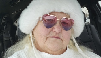 An older woman wearing a plush white fur hat and pink heart-shaped sunglasses inside a car.