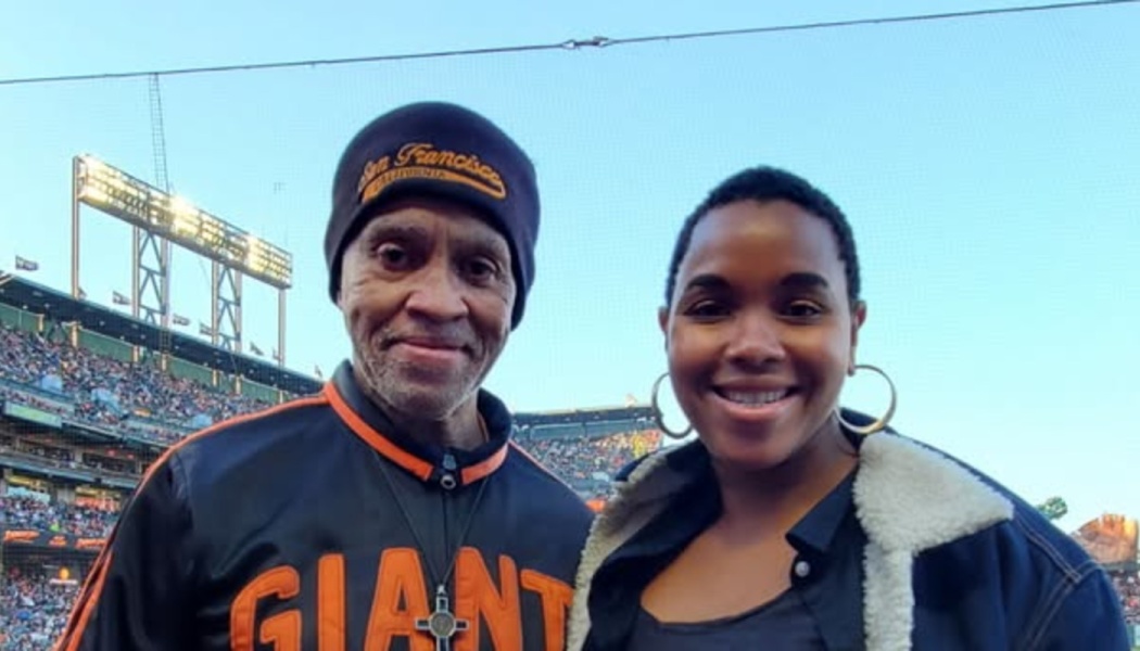 A man in a San Francisco Giants jacket and beanie smiling beside a woman at a baseball stadium.