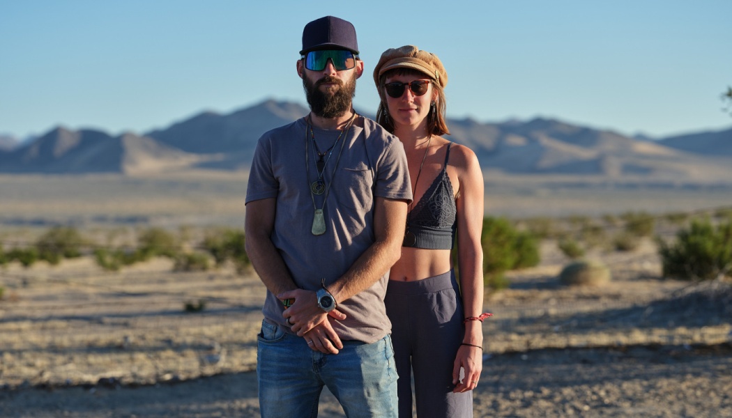 A man and woman wearing sunglasses and hats stand together in a sunny desert with mountains in the background.