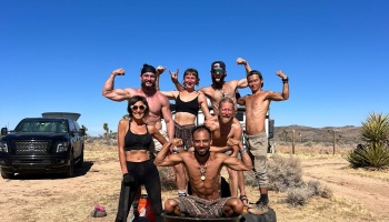 A group of athletic men and women smiling and flexing their muscles in a sunny desert setting.