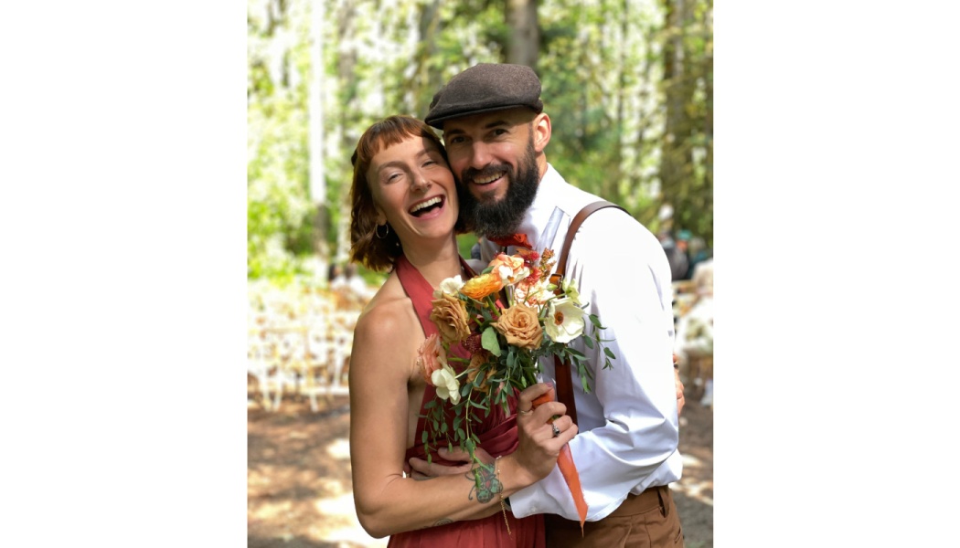 A smiling couple embraces outdoors, the woman holding a bouquet and the bearded man wearing a flat cap and suspenders.