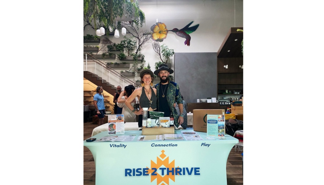 Two founders in hats pose behind a light blue Rise 2 Thrive booth featuring wellness products and flyers.