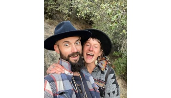 A playful couple wearing wide-brim hats and warm patterned jackets pose closely together outdoors.