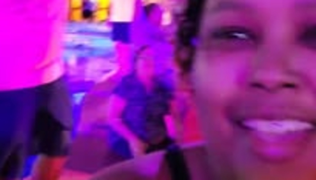 A smiling woman taking a close-up selfie under vibrant pink neon lighting at a crowded indoor event.