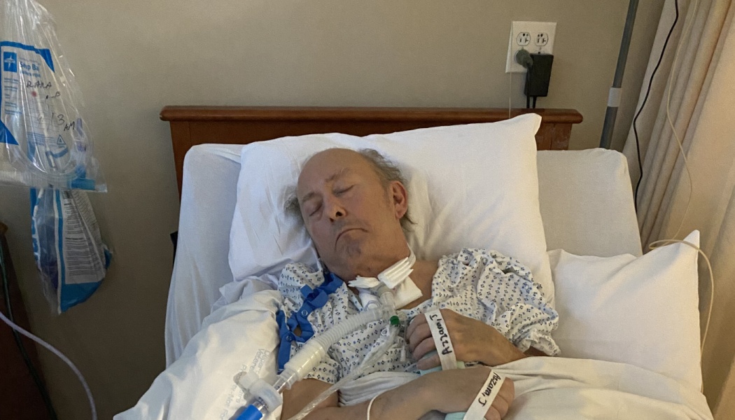 A man resting in a hospital bed with medical tubing and a tracheostomy tube attached.