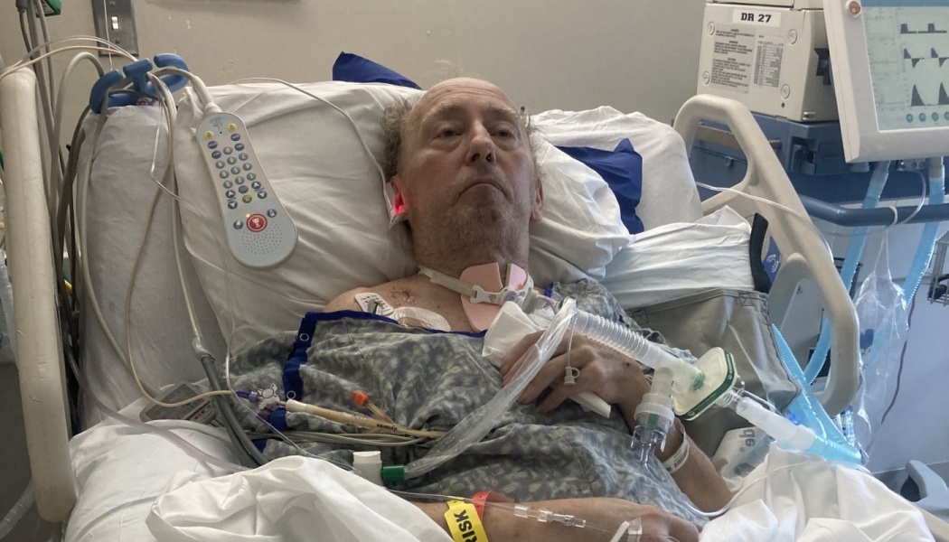An elderly man lying in a hospital bed connected to a tracheostomy tube and medical monitoring equipment.