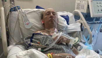 An elderly man lying in a hospital bed connected to a tracheostomy tube and medical monitoring equipment.