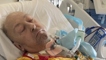 An elderly man lies in a hospital bed with his eyes closed, connected to a ventilator and wearing a neck brace.