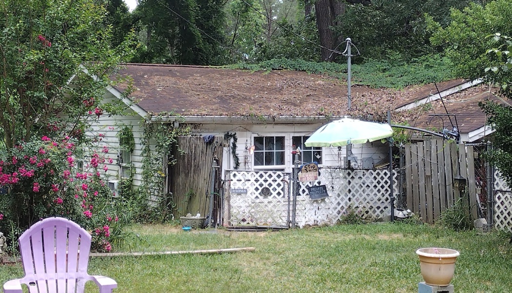 A run-down white home with an aging roof in need of repair, viewed from a grassy yard with a purple lawn chair.