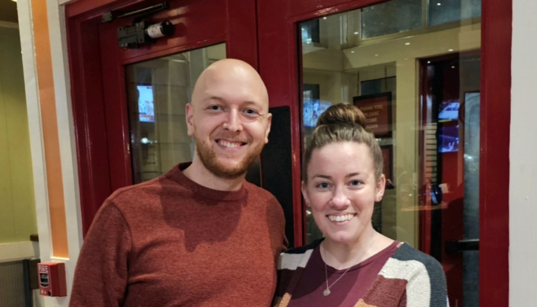 A smiling bald man with a beard and a smiling woman with a top bun pose together in front of red doors.