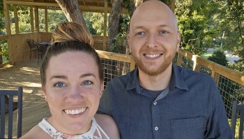 A smiling woman with her hair in a bun and a bald, bearded man taking a selfie on a wooden treehouse deck.