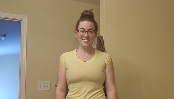 A smiling young woman with glasses and her hair in a high bun, wearing a light yellow t-shirt.
