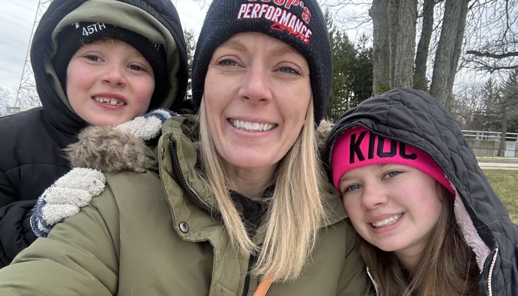 A smiling mother poses outdoors with her son and daughter, all wearing winter beanies and heavy coats.