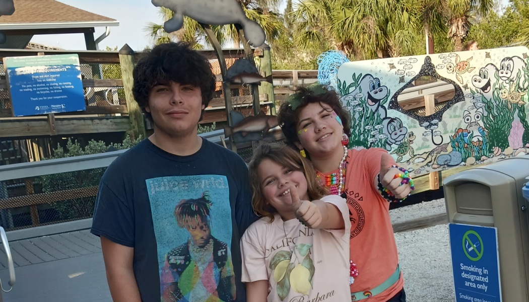 Three smiling young people pose outdoors, two giving thumbs up at a marine life park.