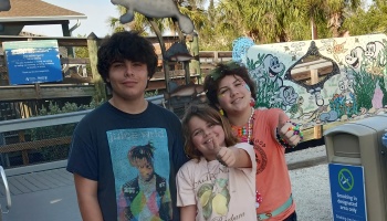 Three smiling young people pose outdoors, two giving thumbs up at a marine life park.