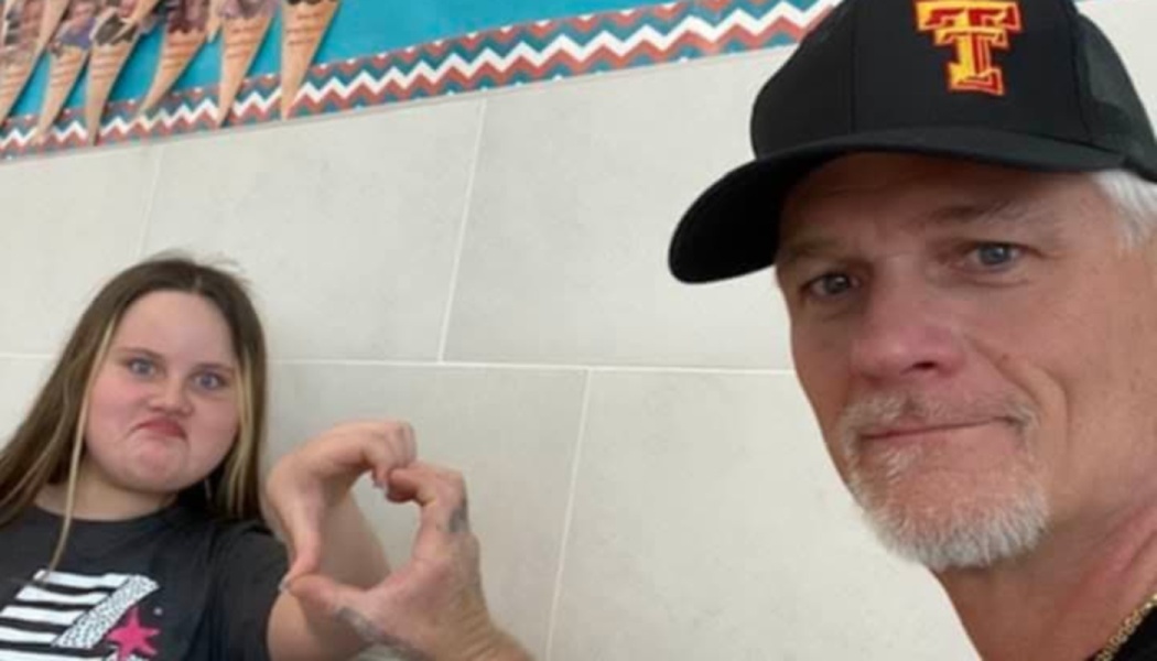A man wearing a baseball cap and a young girl making a funny face form a heart shape together with their hands.