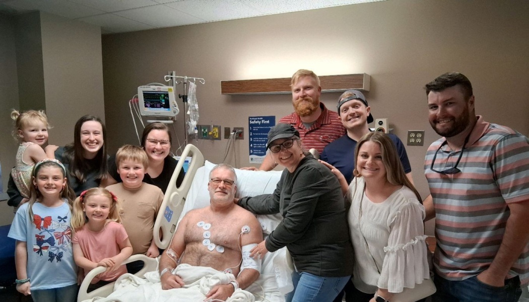 A smiling family with children gathered around a man resting in a hospital bed.