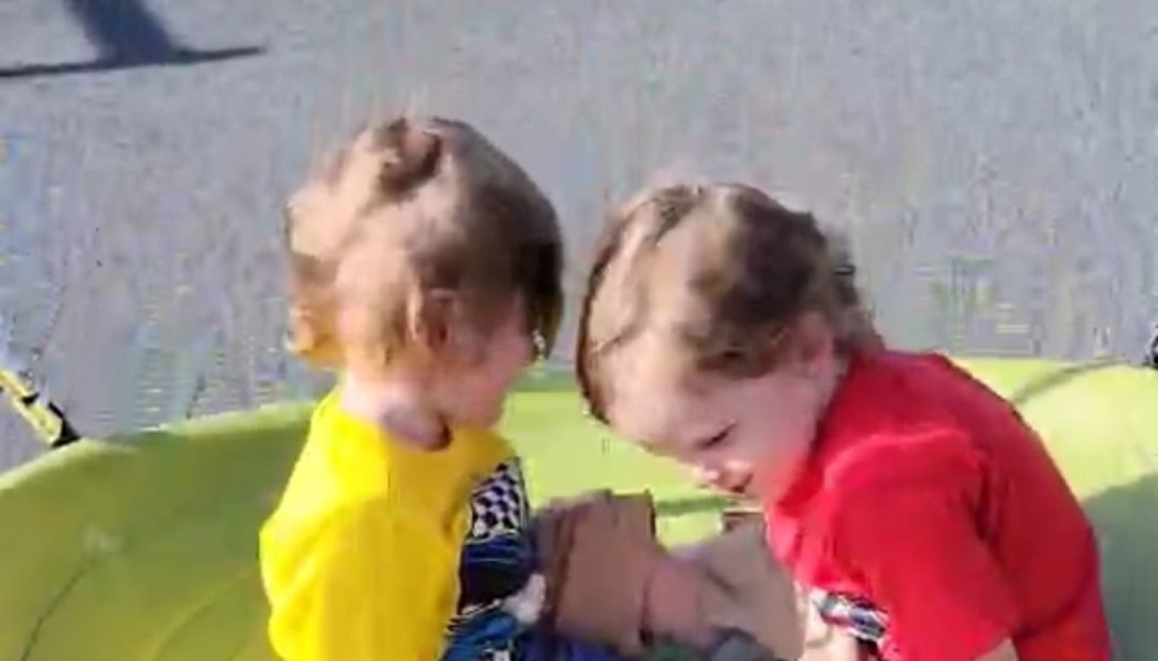 Two young children in yellow and red shirts playing together inside a green round saucer swing outdoors.