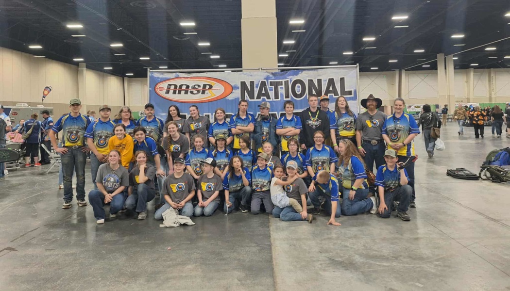 A large youth archery team poses in front of a NASP National Tournament banner at an indoor competition.