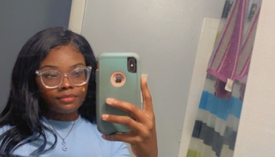 A young Black woman with clear glasses and long dark hair taking a mirror selfie.